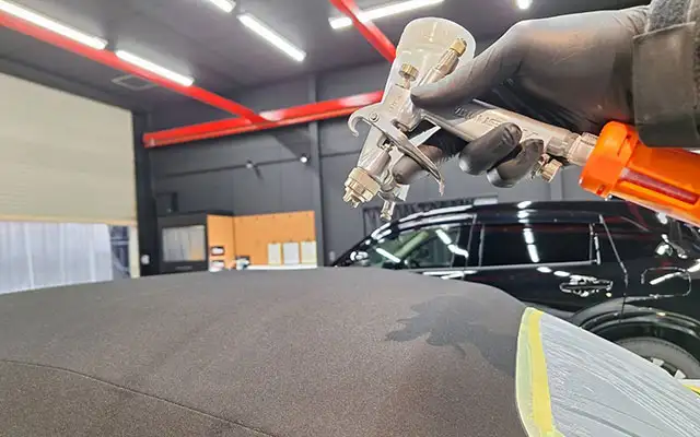 Auto body technician wearing gloves spraying a car roof with a paint gun in a workshop setting