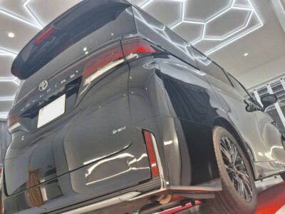 Rear three-quarter view of a dark gray SUV in a showroom with geometric white ceiling lights. License plate obscured for privacy. The vehicle sits on a red carpet next to a glass wall.