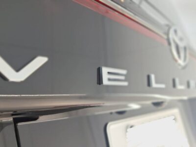 Close-up of a dark car's rear showing large chrome lettering along the trunk and a circular emblem at the right.