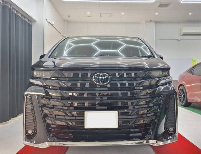 Front view of a glossy black Toyota SUV in a showroom, headlights on, chrome grille and license plate area visible on a red carpet.