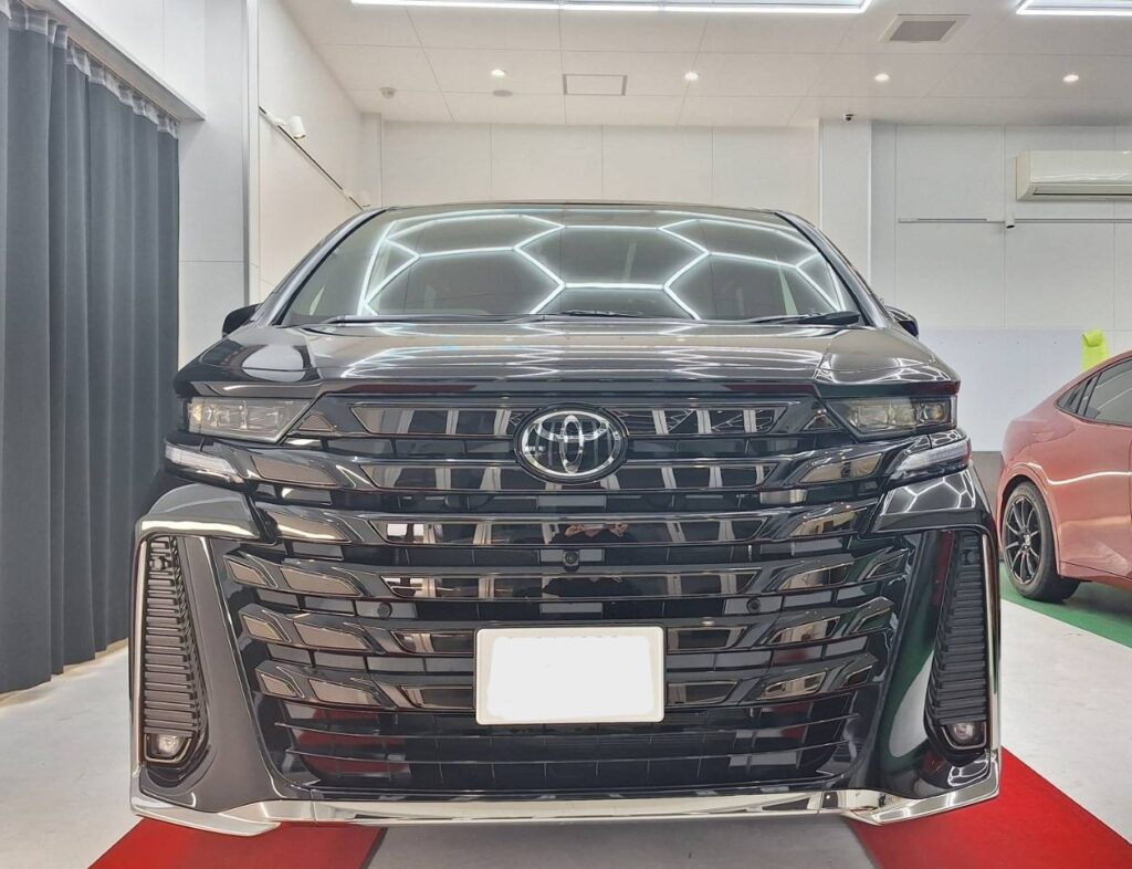 Front view of a glossy black Toyota SUV in a showroom, headlights on, chrome grille and license plate area visible on a red carpet.