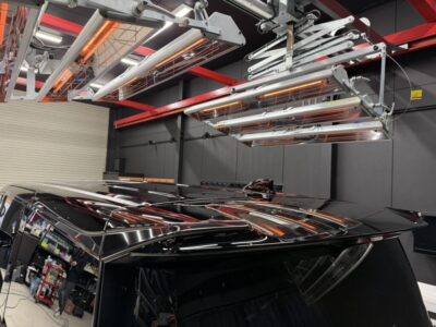 Glossy black SUV in a workshop with ceiling infrared heaters and red support beams overhead