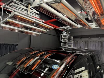 Black SUV under overhead orange infrared heaters in a spray booth/paint booth.
