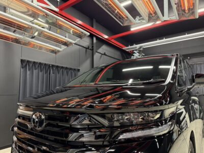 Front view of a glossy black Toyota van on display in a modern showroom, with red structural beams overhead and infrared heaters above.