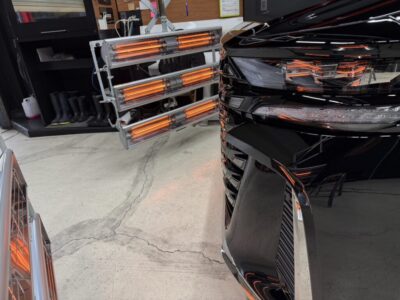 Black car in a workshop next to a rack of orange infrared heaters glowing warmly, with tools and shelves visible in the background.