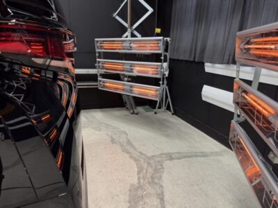 Infrared heat lamps in a car workshop aimed at curing or drying a vehicle’s finish, with a black car on the left and equipment on racks nearby.