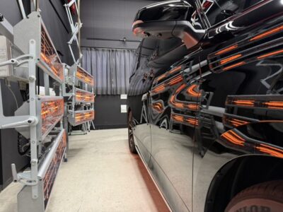 Black SUV parked inside a workshop, reflections visible on its glossy surface from equipment on the left side.