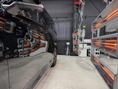 Side view of a black minivan inside an automotive test chamber with orange infrared heater panels on the right and equipment in the background.