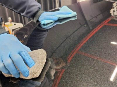 Hands in blue gloves wiping a dark car panel with a microfiber cloth during detailing.