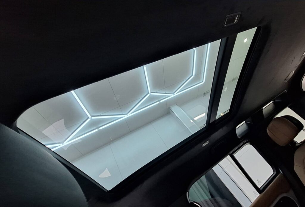 Inside a car, view through the panoramic sunroof at a white ceiling with glowing geometric LED lines outside.