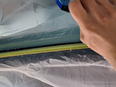 Person in a workshop uses a blue plastic scraper to remove or smooth window film from a glass surface inside a spray booth or workshop.