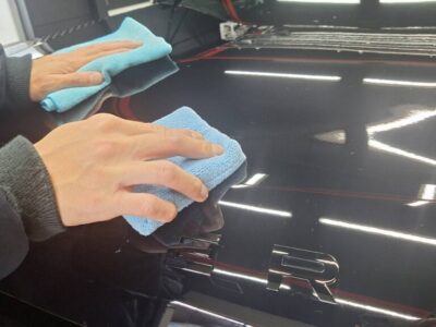 Two hands wiping a glossy black car hood with blue microfiber cloths during detailing, reflections of lights visible on the surface.