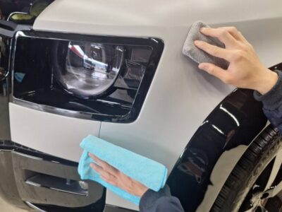 Person polishing the front fender and headlight of a car with microfiber cloths, likely detailing the vehicle surface by hand.
