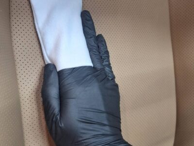 Gloved hand holding a white cloth and resting on a tan perforated car seat, as if cleaning the seat surface.