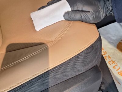 Gloved hand wiping a tan leather car seat with a white cloth inside a vehicle.