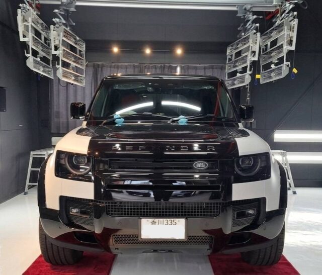 Front view of a two-tone (black and white) Land Rover SUV in a studio setting with stage lighting above and red carpet beneath the vehicle.
