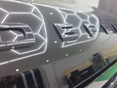 Close-up of a car's chrome badge on a dark, glossy panel with hexagonal light reflections overhead.