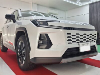 Front three-quarter view of a white SUV in a showroom on a red carpet, angled headlights visible.