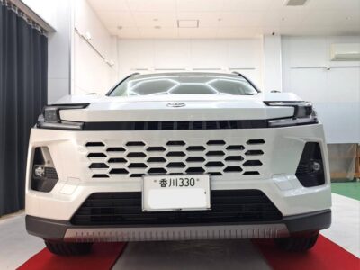 Front view of a white SUV with a large hexagonal grille in a showroom, Japan license plate visible, red carpet beneath.