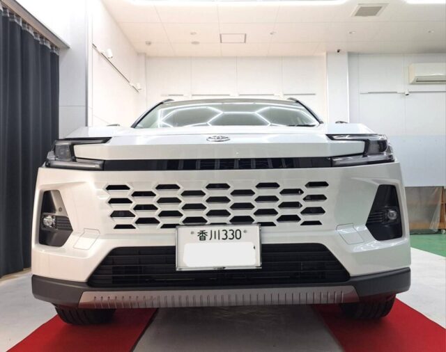 Front view of a white SUV with a large hexagonal grille in a showroom, Japan license plate visible, red carpet beneath.