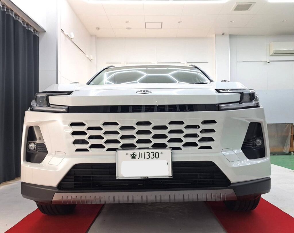 Front view of a white SUV with a large hexagonal grille in a showroom, Japan license plate visible, red carpet beneath.