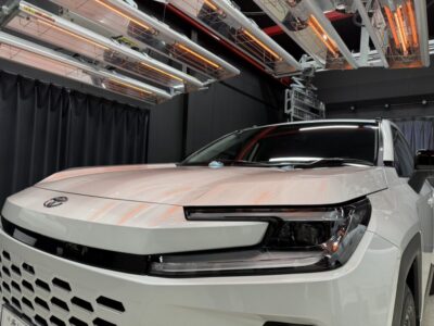 White pickup/SUV in a testing chamber, with orange infrared heater lamps overhead, front view in bright studio lighting.