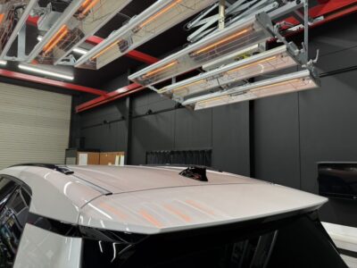White car inside a controlled paint booth with overhead infrared heaters and red supports above.