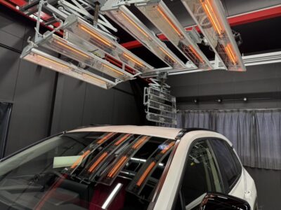 White SUV in a dark testing studio with overhead orange infrared heating lamps and metal racks above the roof