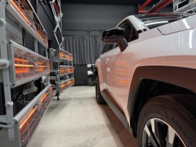 White SUV in a workshop bay beside glowing orange infrared heaters on the wall-mounted racks.