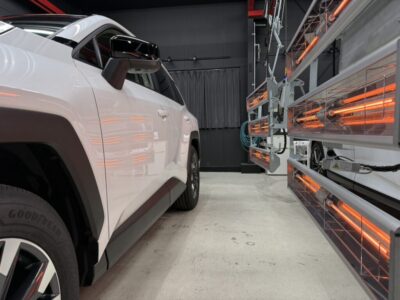 White SUV parked in a car service bay beside orange infrared heater panels along the wall.