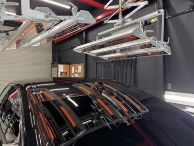 Black car parked in a modern garage under overhead metal rack storage and hoists, with bright shop lights and dark walls in the background.