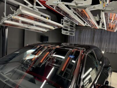Black sedan in a lab-like testing chamber with ceiling-mounted radiant heaters and metal rigging above it.