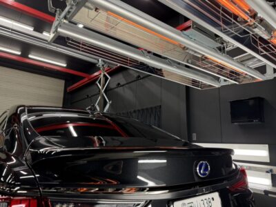 Black Lexus sedan indoors, rear view visible beneath overhead industrial equipment in a dark workshop/garage environment with bright lights and cables overhead