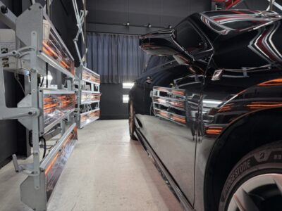 Black car parked in a paint/auto booth with orange infrared curing lamps along the left wall.