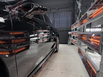 Black car inside a paint-curing booth with orange infrared lamps on the right side.