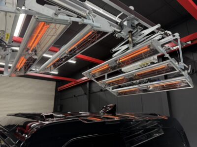 Overhead infrared heating lamps and a metal framework above a glossy black car in a workshop setting.