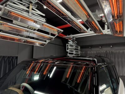 Black SUV inside a lab-like testing chamber with orange infrared heaters and overhead metal racks above. (informative)