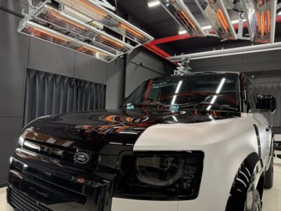 Front three-quarter view of a two-tone Land Rover Defender in black and white, inside a modern showroom with bright overhead lights and gray walls.