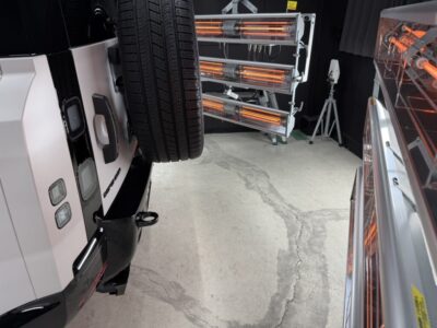 White truck on a lift in a workshop with a large tire mounted to the back and orange infrared heaters in the background.