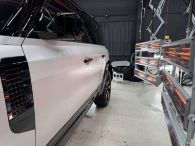 White SUV inside a workshop beside orange infrared curing lamps in use on the right side of the image.
