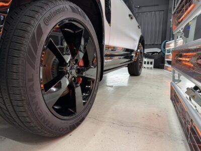 Close-up of a black alloy wheel and Continental tire on a white vehicle in a workshop; red brake calipers visible through the spokes.