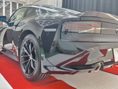 Black sports car shown from the rear-left on a red carpet, glossy finish, distinctive taillights, and large multi‑spoke wheels with a visible exhaust.