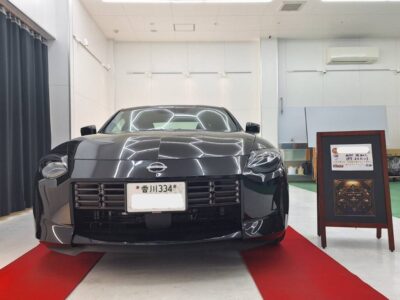 Front view of a black sports car displayed indoors on a red carpet, showroom with a signboard to the right.
