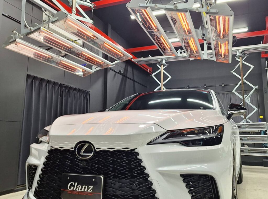 White Lexus sedan under overhead infrared heat lamps in a detailing studio or workshop.