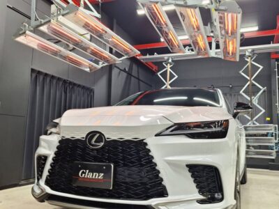 Front view of a white Lexus SUV in a workshop with a large black spindle grille and overhead infrared heaters illuminating the scene nearby.