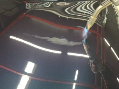 Close-up of a dark blue car hood being welded/heat-treated with a handheld torch in a body shop, creating a blue flame and reflections.