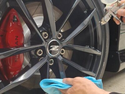 Close-up of a car wheel being cleaned: a red brake caliper, dark alloy rim, and a person wiping the rim with a blue microfiber cloth while a spray bottle is held nearby.