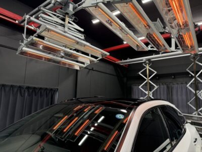 White car in a dark studio with bright orange infrared heating lamps overhead, reflecting on the windshield and roof.