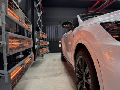 White sports car parked in a workshop with orange infrared heater racks on the left side.