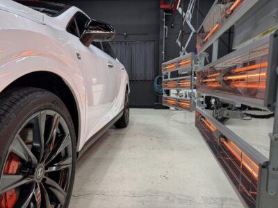 White luxury SUV in a workshop beside orange infrared curing lamps used for paint curing/finishing.